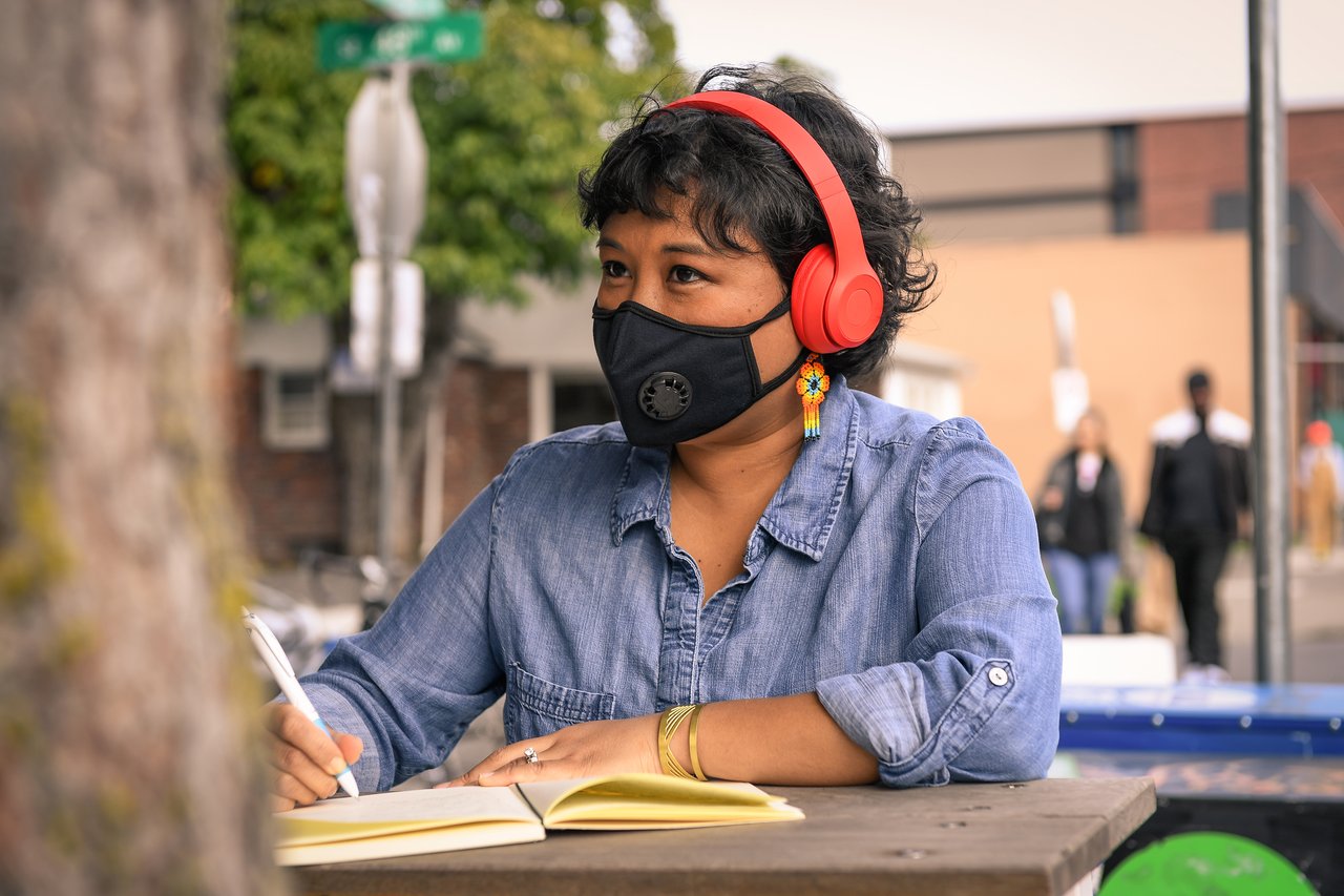 A woman in red headphones and a face mask writing in a notebook at an outdoor table