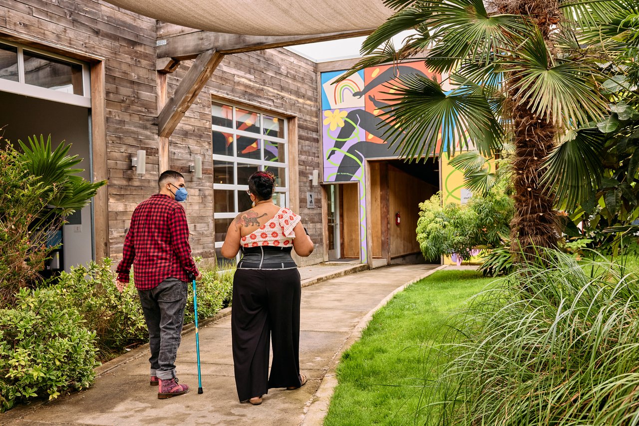 Two people walking together through a garden courtyard, one using a cane