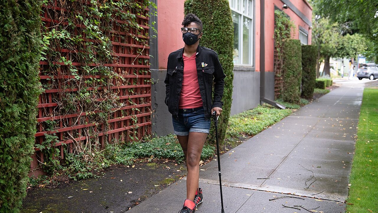 A person wearing a face mask walks with a cane along a tree-lined neighborhood sidewalk