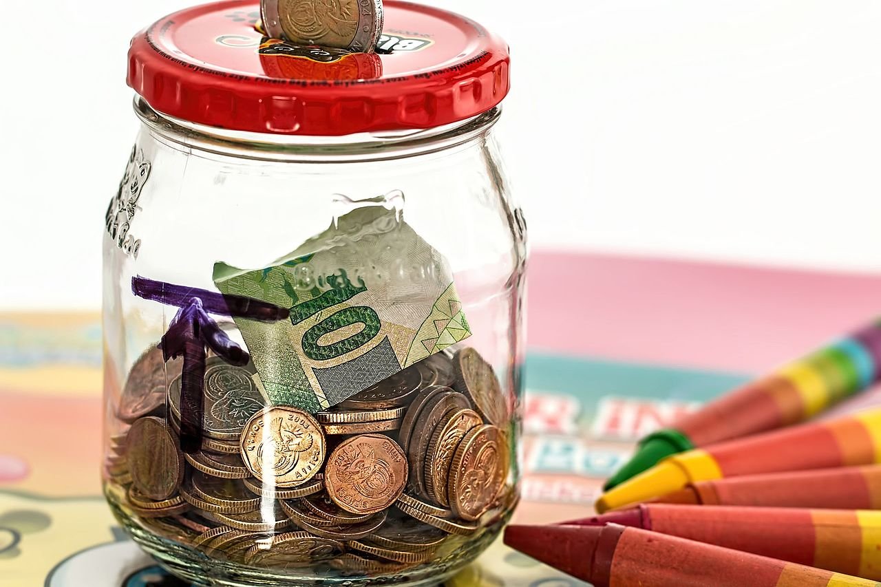 A glass savings jar holding coins and a folded bill next to crayons