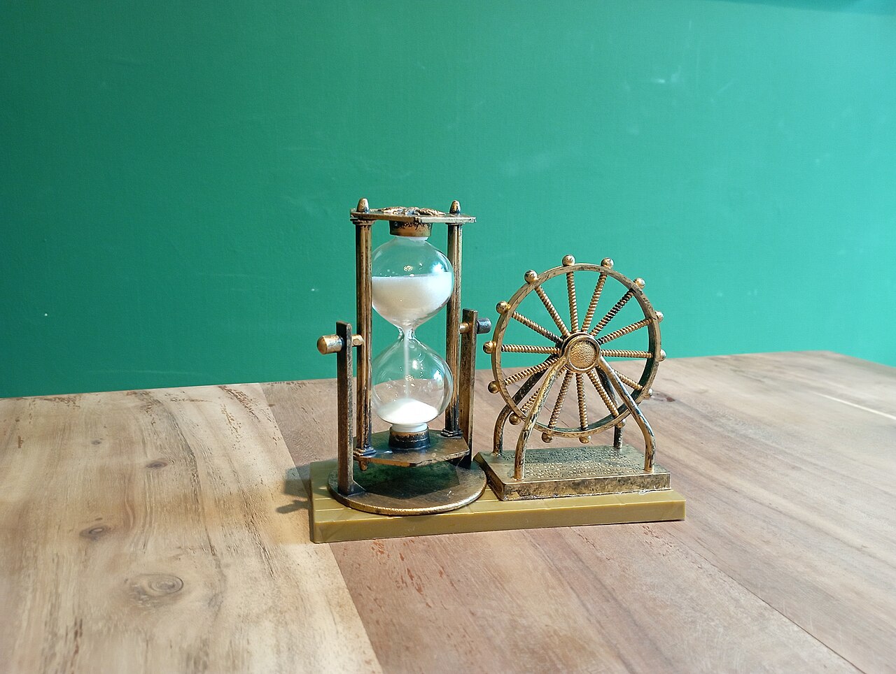 A brass hourglass with white sand beside a small decorative wheel on a wooden table
