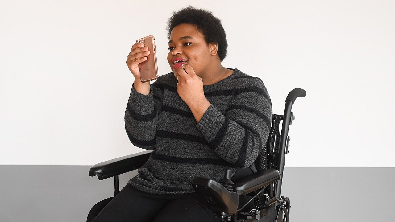 A person seated in a power wheelchair applies lip gloss, using a phone as a mirror