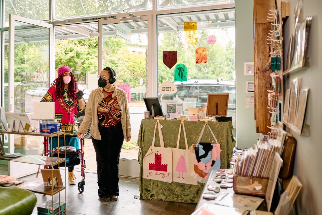 Two disabled friends in face masks walk into a gift shop, one using a rollator