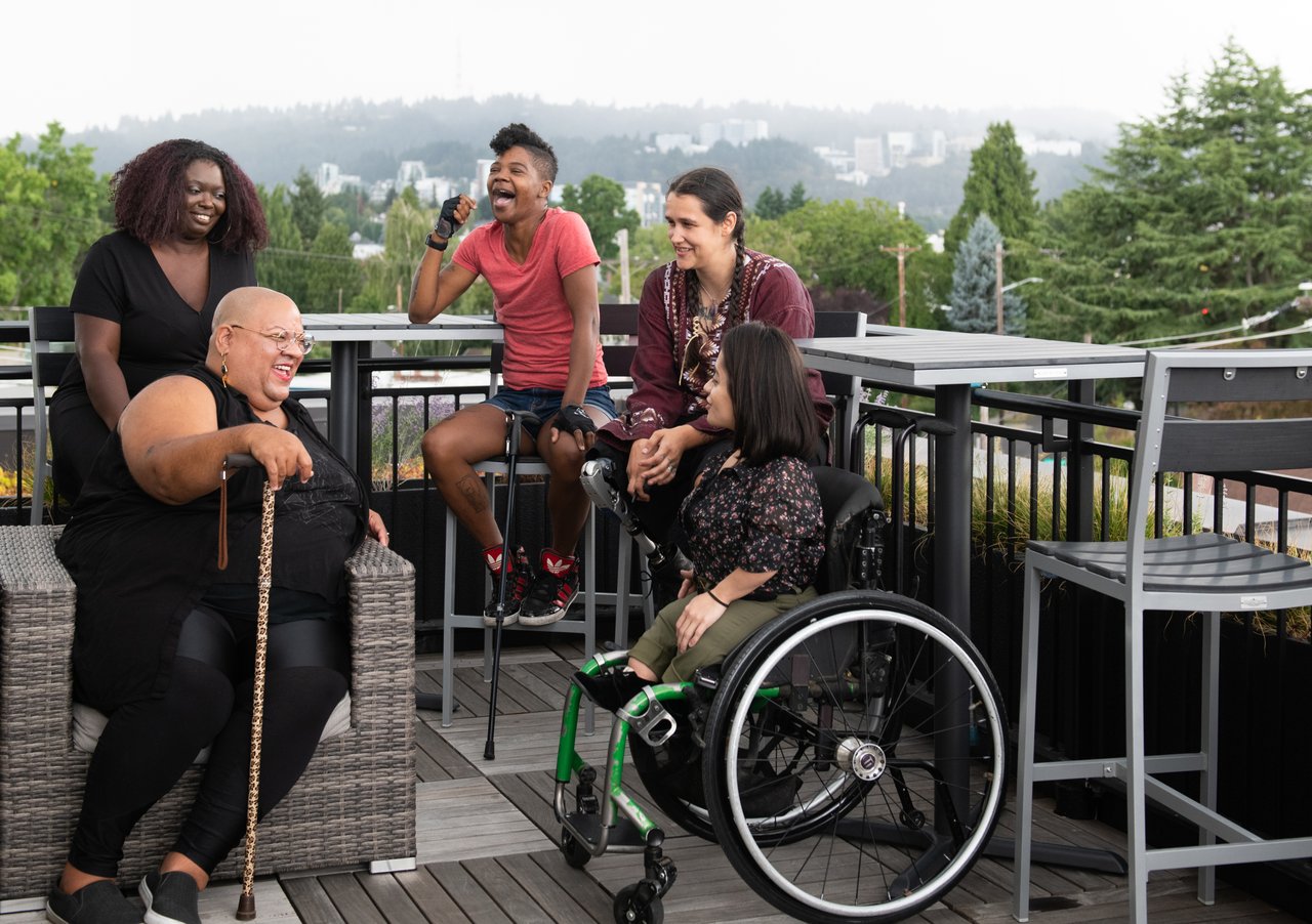 Five friends with canes and a wheelchair laughing together on a rooftop deck