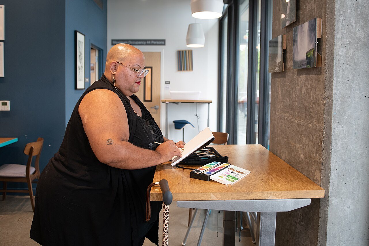 A Black non-binary person paints at an adjustable desk