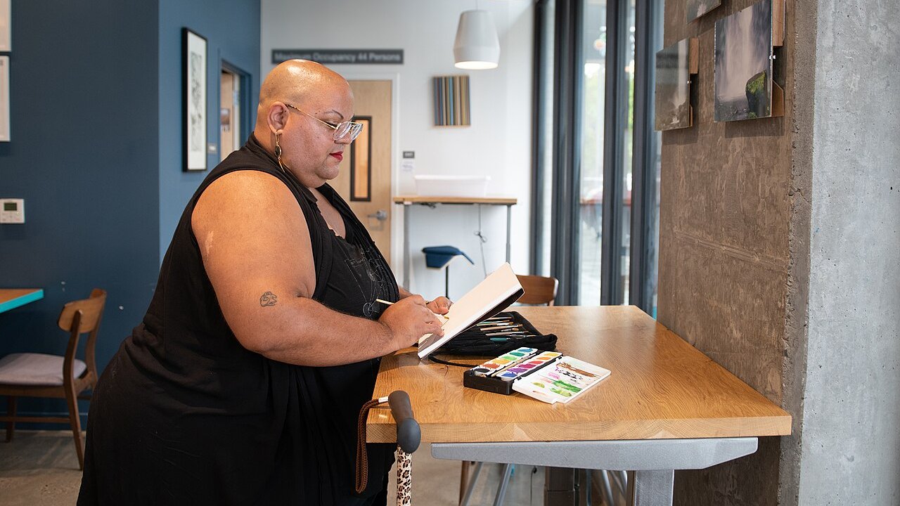 A person in black with a leopard-print cane hanging from an adjustable desk paints with watercolors in a cafe