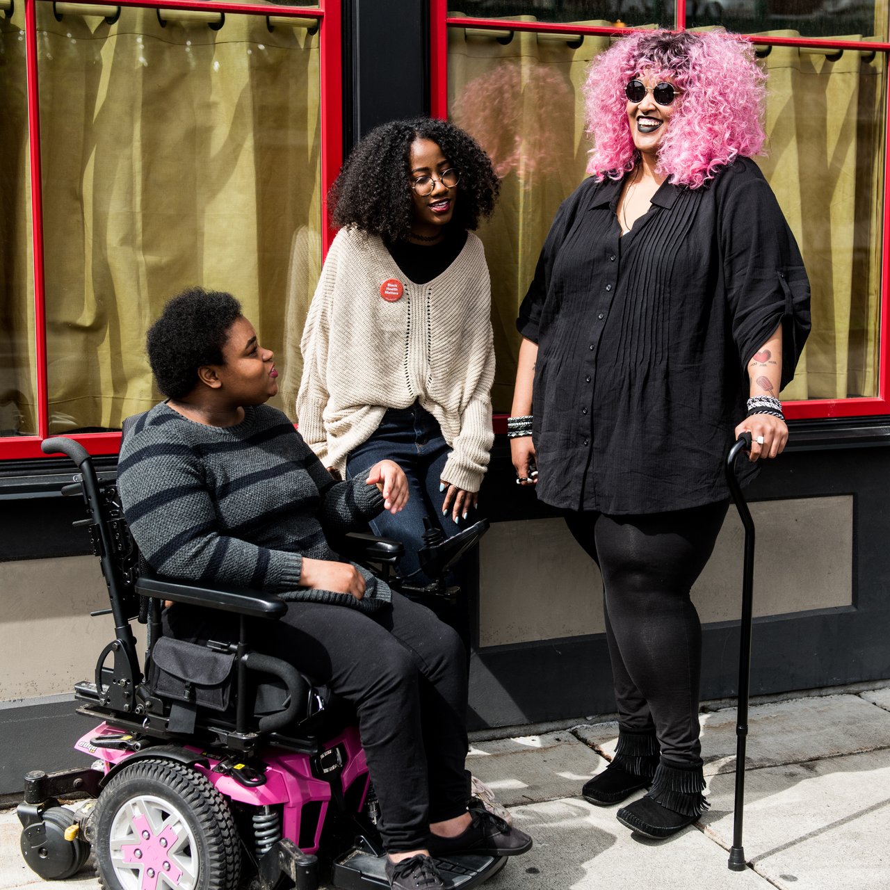 Three friends talking outside a building, one using a power wheelchair
