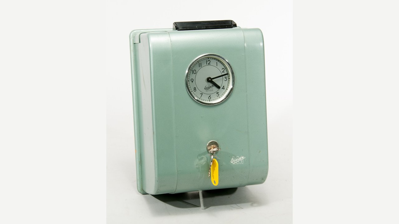 A green 1950s punch time clock with a round clock face and a key in its lock